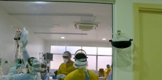 profissionais de saúde utilizando EPI em volta de paciente em cama vistos por grande janela de vidro em corredor