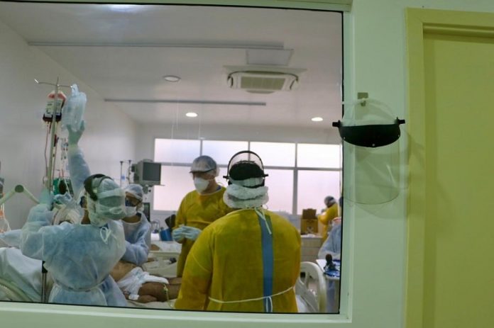 profissionais de saúde utilizando EPI em volta de paciente em cama vistos por grande janela de vidro em corredor