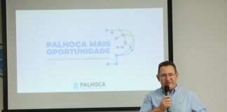 O prefeito Eduardo Freccia é branco, tem cabelo escuros e curtos, usa uma camisa azul clara e segura um microfone. Ao fundo, um telão mostra: Palhoça Mais Oportunidade, progrrama de retomada ecônomica e social