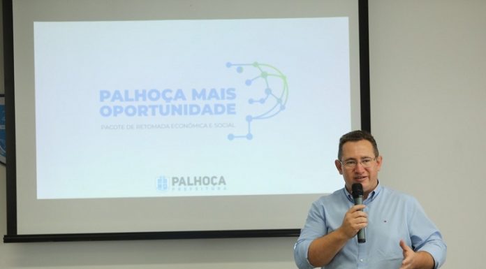 O prefeito Eduardo Freccia é branco, tem cabelo escuros e curtos, usa uma camisa azul clara e segura um microfone. Ao fundo, um telão mostra: Palhoça Mais Oportunidade, progrrama de retomada ecônomica e social