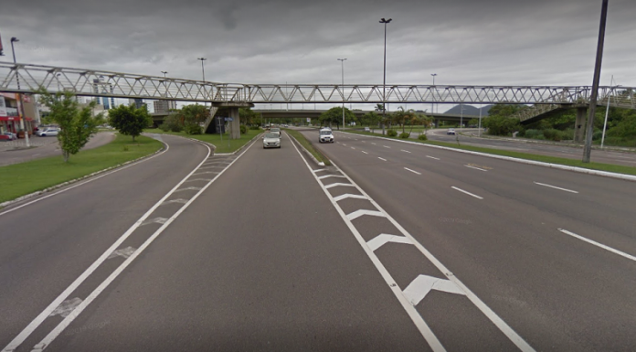 Na avenida, o asfalto cinza com marcações em branco, ao fundo a passarela do CIC, que deve passar por recuperação, de acordo com recomendação do MPSC