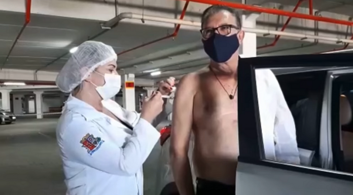 Uma profissional da saúde usa toca, máscara e jaleco branco com o brasão de São José. Ela aplica a vacina contra a covid no prefeito Orvino, que usa máscara escura, colar e está com a camisa em um só braço, parado em pé ao lado do carro.