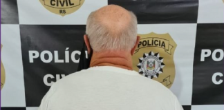 Em frente ao banner com quadrados pretos e brancos que identificam a polícia civil, um homem branco de cabelos grisalhos aparece de costas para a foto com as mãos algemadas para trás. O homem é acusado pelo feminicídio da então companheira, em Itapema.