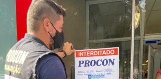 Um fiscal com colete do Procon, acompanhado do secretário Gabrielzinho, colam na porta da agência bancária um papel escrito "interditado" e "Procon". Ambos usam máscara.