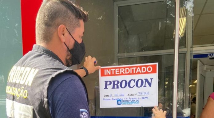 Um fiscal com colete do Procon, acompanhado do secretário Gabrielzinho, colam na porta da agência bancária um papel escrito "interditado" e "Procon". Ambos usam máscara.