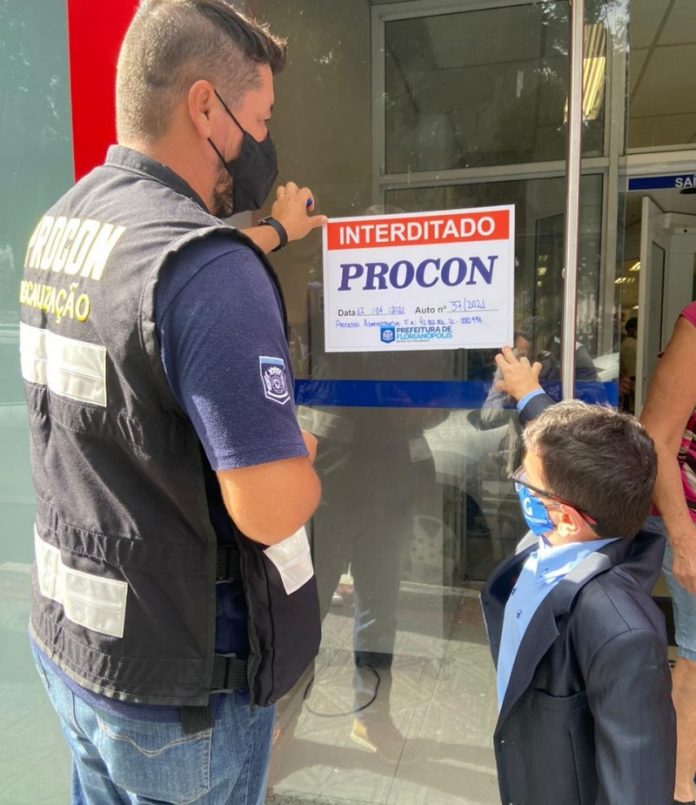 Um fiscal com colete do Procon, acompanhado do secretário Gabrielzinho, colam na porta da agência bancária um papel escrito 