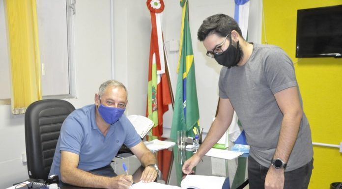 O prefeito de Governador Celso Ramos assina o convênio que destina recursos para a reconstrução de estruturas esportivas da cidade, destruídas pelo ciclone bomba de 2020. Ele está sentado usando máscara e camiseta azuis, ele é branco e tem cabeços grisalhos. Do outro lado da mesa, um rapaz de máscara preta, pele branca cabelos escuros e de óculos, acompanha a assinatura.