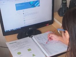 Alunos da rede estadual ainda não iniciaram as aulas no ensino remoto tela do computador com algumas atividades e abaixo um caderno com anotações, uma menina aparece no canto, ela está de costas e usa óculos, ela está realizando as atividades escolares. A foto representa o ensino remoto.