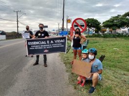 No acostamento de uma rua, grevistas seguram cartazes em que é possível ler "essencial é a vida". Três pessoas estão em pé e uma agachada com uma criança. A greve dos servidores da educação municipal de Florianópolis segue sem acordo com a prefeitura.