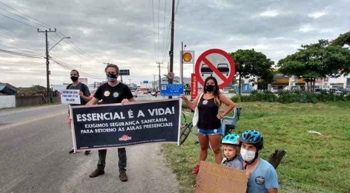 No acostamento de uma rua, grevistas seguram cartazes em que é possível ler "essencial é a vida". Três pessoas estão em pé e uma agachada com uma criança. A greve dos servidores da educação municipal de Florianópolis segue sem acordo com a prefeitura.