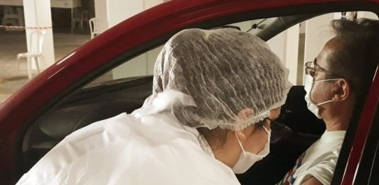 São José inicia vacinação contra a Covid-19 para idosos de 64 e 63 anos mulher vestida de branco, com touca na cabeça, aplicando vacina no braço de homem sentado dentro de um carro vermelho