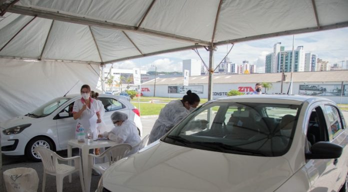 dois carros parados para vacinação sob tenda de drive-thru com profissionais de saúde em volta