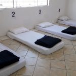 Em um quarto branco com janela, quatro colchões estão encostados na parece com numerações acima, a roupa de cama é branca e o cobertor dobrado é escuro. O abrigo emergencial de São José foi montado para atender as pessoas durante as noites de frio intenso.