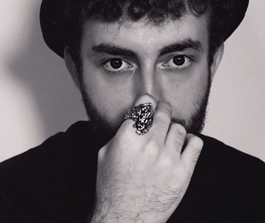 Cantor e compositor catarinense Assini lança o single “dancing alone” Em foto preto e branco o cantor Assini aparece com chapéu escuro e blusa escura, ele tem barba e alguns cachos de cabelo na testa, sua mão está na frente de seu rosto com um anel no dedo indicador