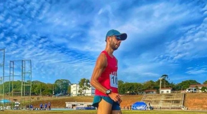 Um atleta de camiseta vermelha, tênis, boné e shorts azuis, além de óculos escuros corre em marcha atlética em uma pista de cor azul. O time de atletismo de São José conquistou vaga nos jogos sul-americanos