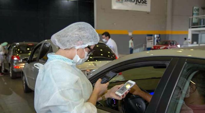 Prefeituras ampliam vacinação de pessoas com comorbidades; veja as idades homem mostra dentro de carro celular com qr-code à funcionária de vacinação dentro de ginásio - vacinação de pessoas com comorbidades