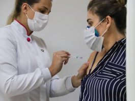 Uma mulher de jaleco e mascaras brancas, usa o cabelo preso em rabo de cavalo e aplica a vacina em outra mulher, de cabelo preso, máscara e blusa listrada. As pessoas com comorbidades de 55 a 57 anos serão vacinadas na próxima segunda em Florianópolis.