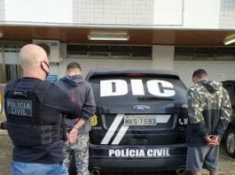 policial careca olha para os dois jovens presos com as mãos algemadas atrás de uma viatura, todos estão de costas - Homens que mataram motorista de aplicativo em São José são presos