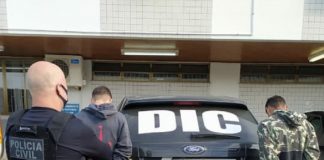 policial careca olha para os dois jovens presos com as mãos algemadas atrás de uma viatura, todos estão de costas - Homens que mataram motorista de aplicativo em São José são presos