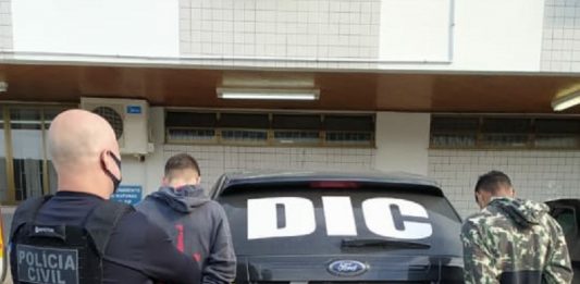 policial careca olha para os dois jovens presos com as mãos algemadas atrás de uma viatura, todos estão de costas - Homens que mataram motorista de aplicativo em São José são presos