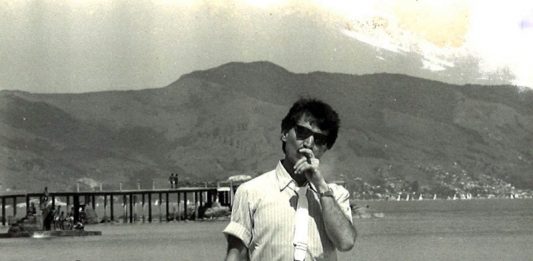 belchior na praia da saude em coqueiros, fumando um cigarro e de óculos escuros; ao fundo pessoas no trapiche