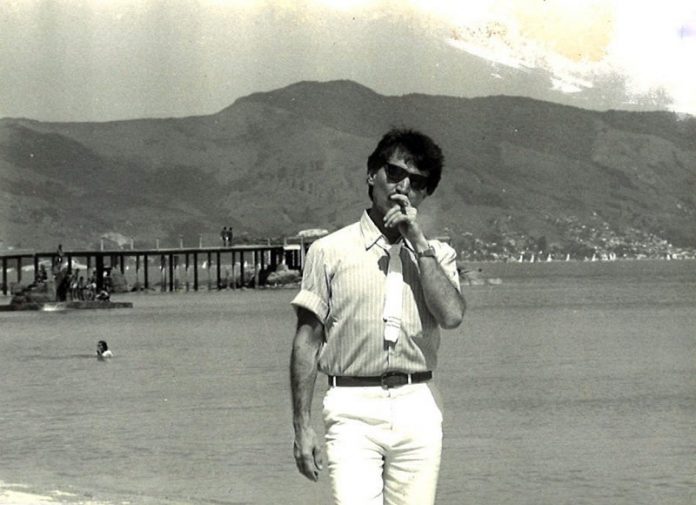 belchior na praia da saude em coqueiros, fumando um cigarro e de óculos escuros; ao fundo pessoas no trapiche