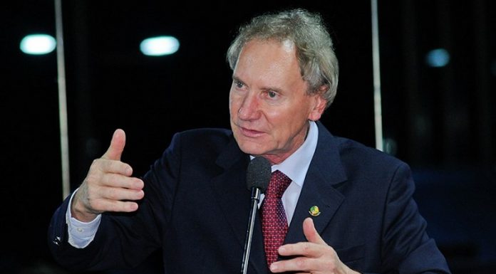 Em registro do senado, Casildo Maldaner aparece de cabelos brancos, pele branca, terno escuro e gravata vermelha, gesticulando com as mãos em frente a um microfone