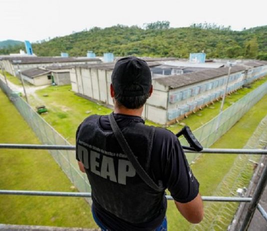 De costas, uma pessoa aparece de boné, camiseta e colete pretos, ele carrega um armamento e observa uma área que parece ser um presídio. Nesta semana, o governo convocou 213 policias penais