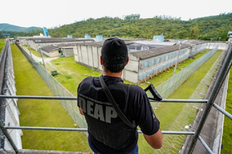 De costas, uma pessoa aparece de boné, camiseta e colete pretos, ele carrega um armamento e observa uma área que parece ser um presídio. Nesta semana, o governo convocou 213 policias penais