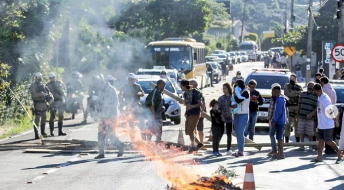 Na imagem um grupo de pessoas protesta, há um ponto de fogo em toras na pista e uma fila de carros. Os moradores do Morro das Pedras protestaram por resoluções para o problema de destruição de imóveis na beira da praia