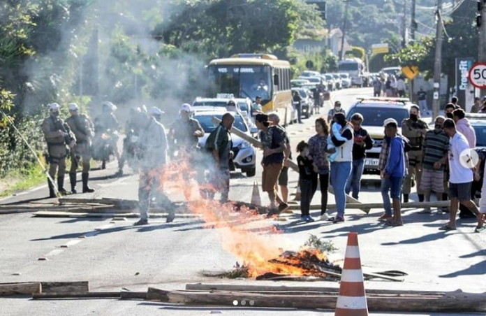 Na imagem um grupo de pessoas protesta, há um ponto de fogo em toras na pista e uma fila de carros. Os moradores do Morro das Pedras protestaram por resoluções para o problema de destruição de imóveis na beira da praia