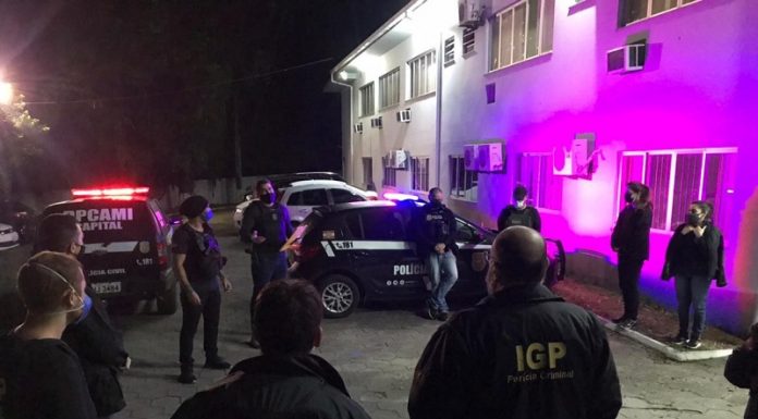 Policiais civis estão em circulo e uniformizados, no cenário ainda aparecem carros da polícia, eles cumpriram mandados e prenderam dois homens na operação de combate à pornografia infantojuvenil em Florianópolis