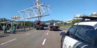 No canto direito um carro da guarda municipal de Florianópolis, ao centro uma parte da passarela suspensa e dois caminhões abaixo. No canto esquerdo outra parte da estrutura no chão e dois funcionários de camisetas verdes.