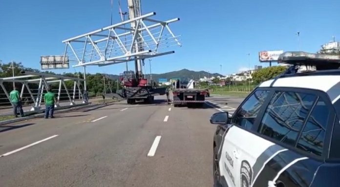 No canto direito um carro da guarda municipal de Florianópolis, ao centro uma parte da passarela suspensa e dois caminhões abaixo. No canto esquerdo outra parte da estrutura no chão e dois funcionários de camisetas verdes.