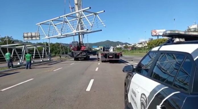 No canto direito um carro da guarda municipal de Florianópolis, ao centro uma parte da passarela suspensa e dois caminhões abaixo. No canto esquerdo outra parte da estrutura no chão e dois funcionários de camisetas verdes.