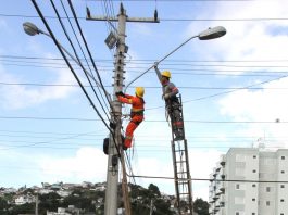 O céu azul e no alto de um poste dois funcionários mexem na iluminação e fios do poste. No Kobrasol, as ruas vão receber a iluminação em led.