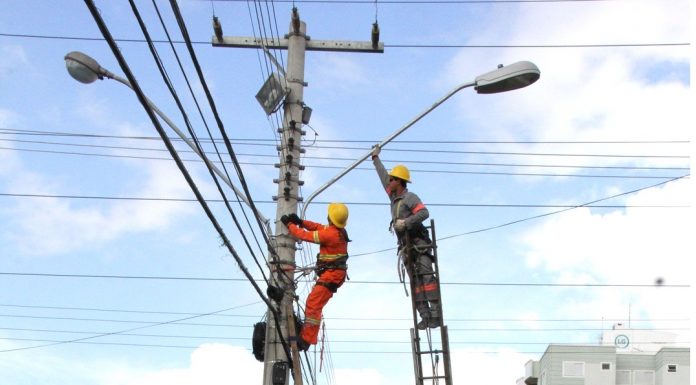 O céu azul e no alto de um poste dois funcionários mexem na iluminação e fios do poste. No Kobrasol, as ruas vão receber a iluminação em led.