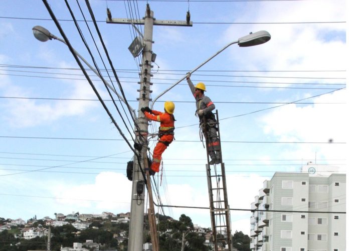 O céu azul e no alto de um poste dois funcionários mexem na iluminação e fios do poste. No Kobrasol, as ruas vão receber a iluminação em led.