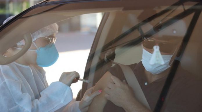 Através do vidro da frente de um carro é possível ver um profissional de saúde com máscara e proteção na cabeça e roupas aplicando a vacina em um senhor que está dentro do carro usando máscara e óculos escuros. Nesta semana, Florianópolis segue a vacinação contra a gripe.