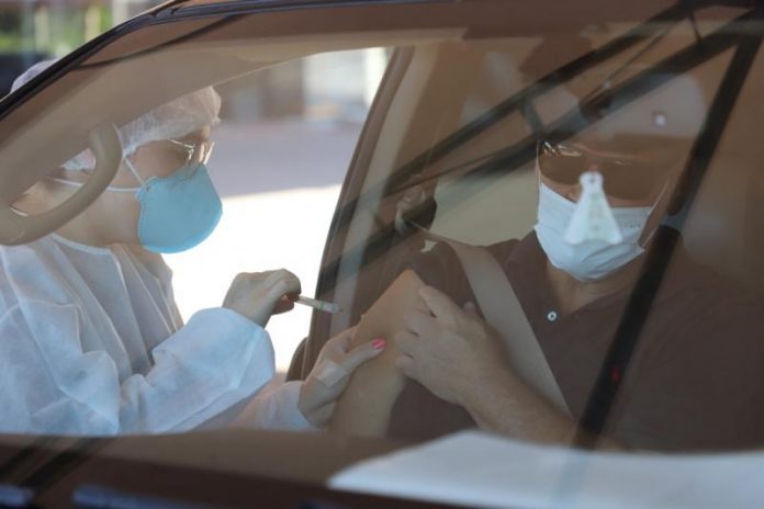 Através do vidro da frente de um carro é possível ver um profissional de saúde com máscara e proteção na cabeça e roupas aplicando a vacina em um senhor que está dentro do carro usando máscara e óculos escuros. Nesta semana, Florianópolis segue a vacinação contra a gripe.