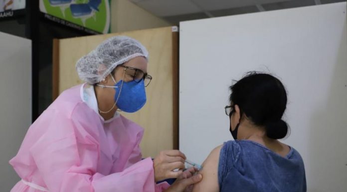 Profissional de saúde com proteção rosa no corspo, usa proteção também no cabelo e usa máscara azul, ela aplica a vacina em uma mulher sentada de costas, com blusa azul. Nesta semana, Florianópolis segue a vacinação de pessoas com comorbidades maiores de 18 anos e pessoas acima dos 60.