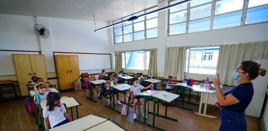 professora usando máscara dá aula para crianças em carteiras usando máscaras - doses para vacinação de professores