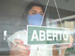 mulher com roupa branca e máscara azul colocando uma placa de aberto na porta do estabelecimento