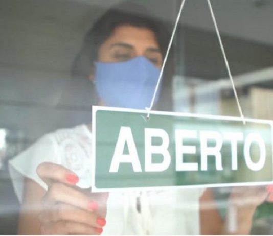 mulher com roupa branca e máscara azul colocando uma placa de aberto na porta do estabelecimento
