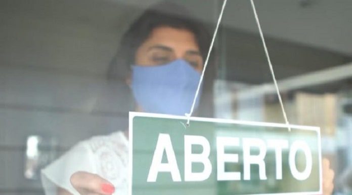 mulher com roupa branca e máscara azul colocando uma placa de aberto na porta do estabelecimento