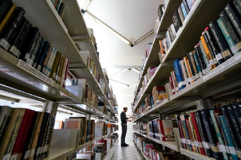 Entre duas estantes com livros em uma biblioteca um homem aparece ao fundo mexendo nos livros. A foto representa o anúncio de bolsas de pós-graduação em Santa Catarina