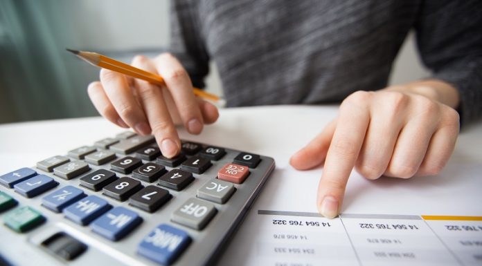 uma mão de cor branca segura um lápis amarelo enquanto digits em uma calculadora, a outra mão aponta para um número em um papel. O Pronampe voltou, mas com juros maiores