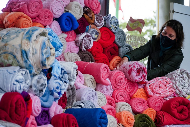 Cobertores coloridos enrolados e empilhados, ao fundo uma mulher de preto os arruma. Eles fazem parte da campanha de doação cobertor solidário.
