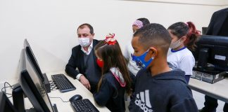 A foto mostra o governador Carlos Moisés sentado em frente a um computador, ele utiliza máscara e está acompanhado de cinco crianças, também de máscara. Cinco escolas da região devem receber laboratórios de informática, escolas de 13 municípios receberão equipamentos.