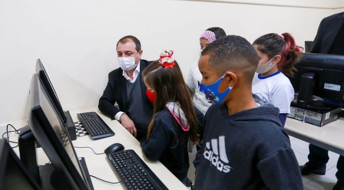 A foto mostra o governador Carlos Moisés sentado em frente a um computador, ele utiliza máscara e está acompanhado de cinco crianças, também de máscara. Cinco escolas da região devem receber laboratórios de informática, escolas de 13 municípios receberão equipamentos.